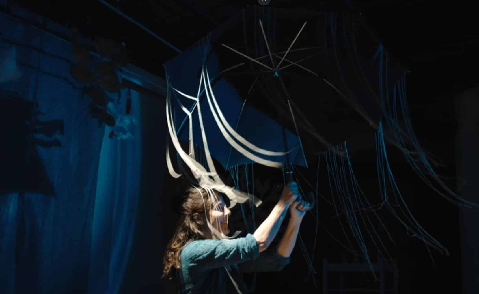 Visuel du spectacle Ah l’eau ! : une interprète manipule un grand parapluie noir entouré de rubans éclairés en bleu.