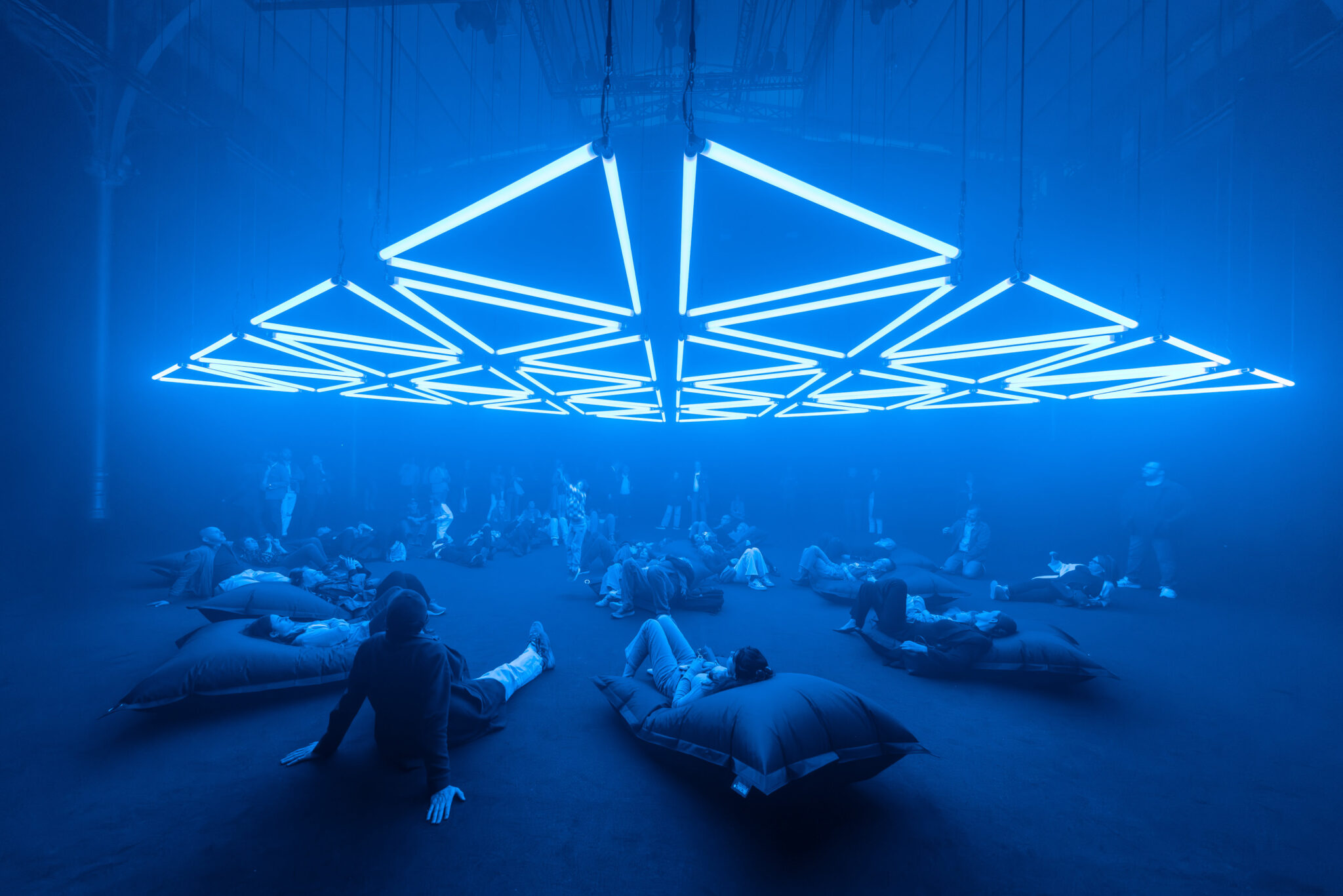 Personnes allongées au sol contemplant une oeuvre lumineuse faite de néons bleus qui forment des triangles et des formes géometriques