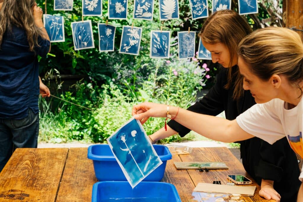 Atelier cyanotype aux Jardins Passagers : des participantes réalisent des impressions bleues de végétaux en les rinçant dans des bacs d’eau, tandis que des œuvres sèchent sur des fils.