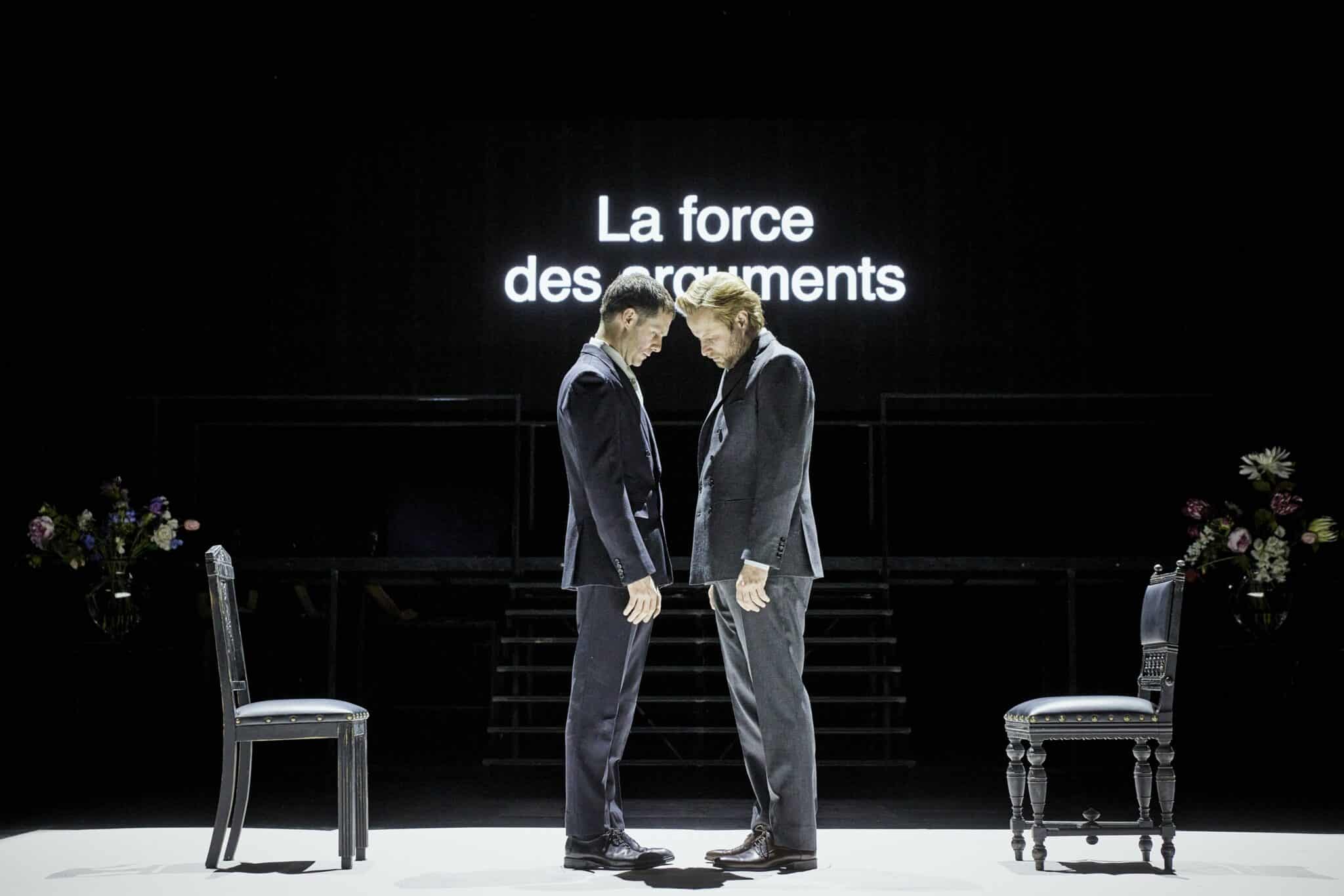 Deux interprètes du Tartuffe ou l’Hypocrite se font face en silence sur un plateau épuré, devant l’inscription lumineuse « La force des arguments ».