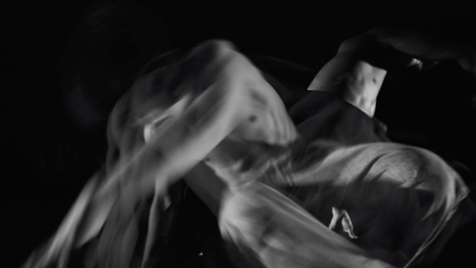 Image en noir et blanc montrant un corps humain partiellement nu en mouvement, créant un effet flou artistique abstrait.
