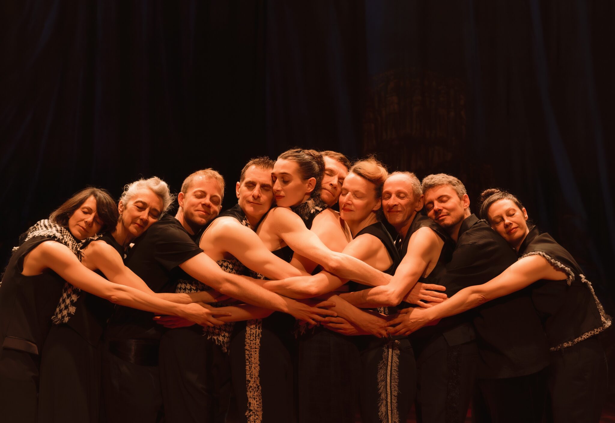 Dix danseurs sont debout en cercle très serré, bras croisés autour des épaules, formant un nœud humain. Ils sont habillés de noir, pieds nus sur un sol en bois. Rideau sombre en fond.