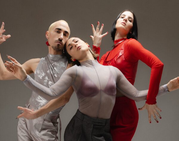Trois danseurs et danseuses dans une salle vide, regardant la caméra, éclairés par une lumière rasante