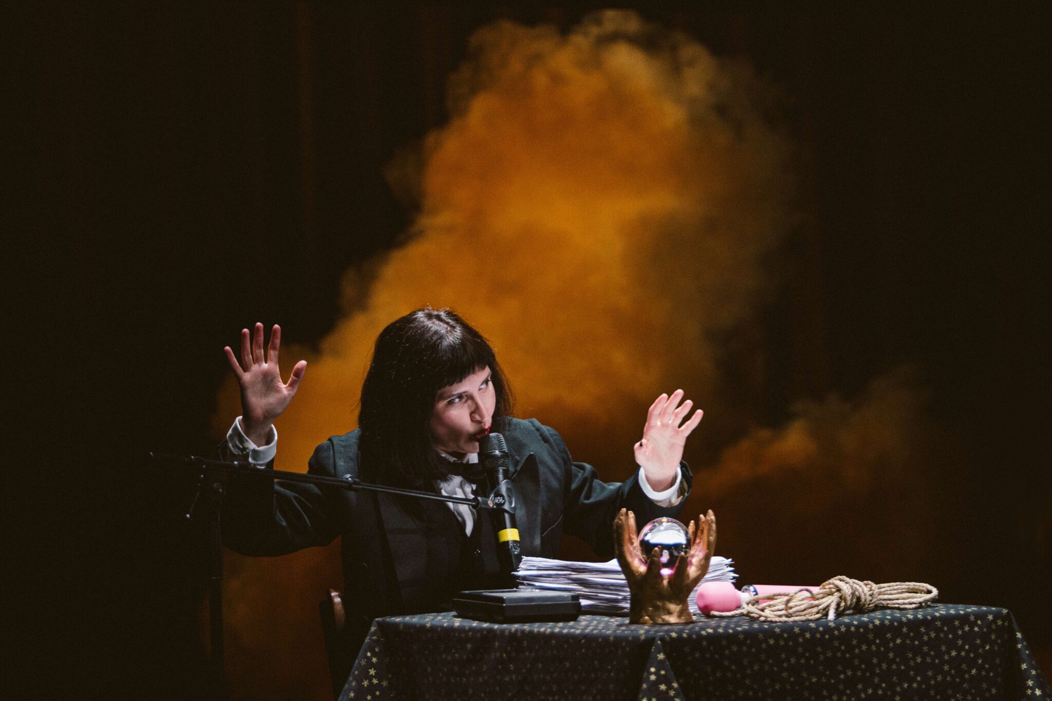 Performeuse en costume à une table recouverte d'étoiles, en pleine action sur scène avec micro, accessoires et fond de fumée orangée