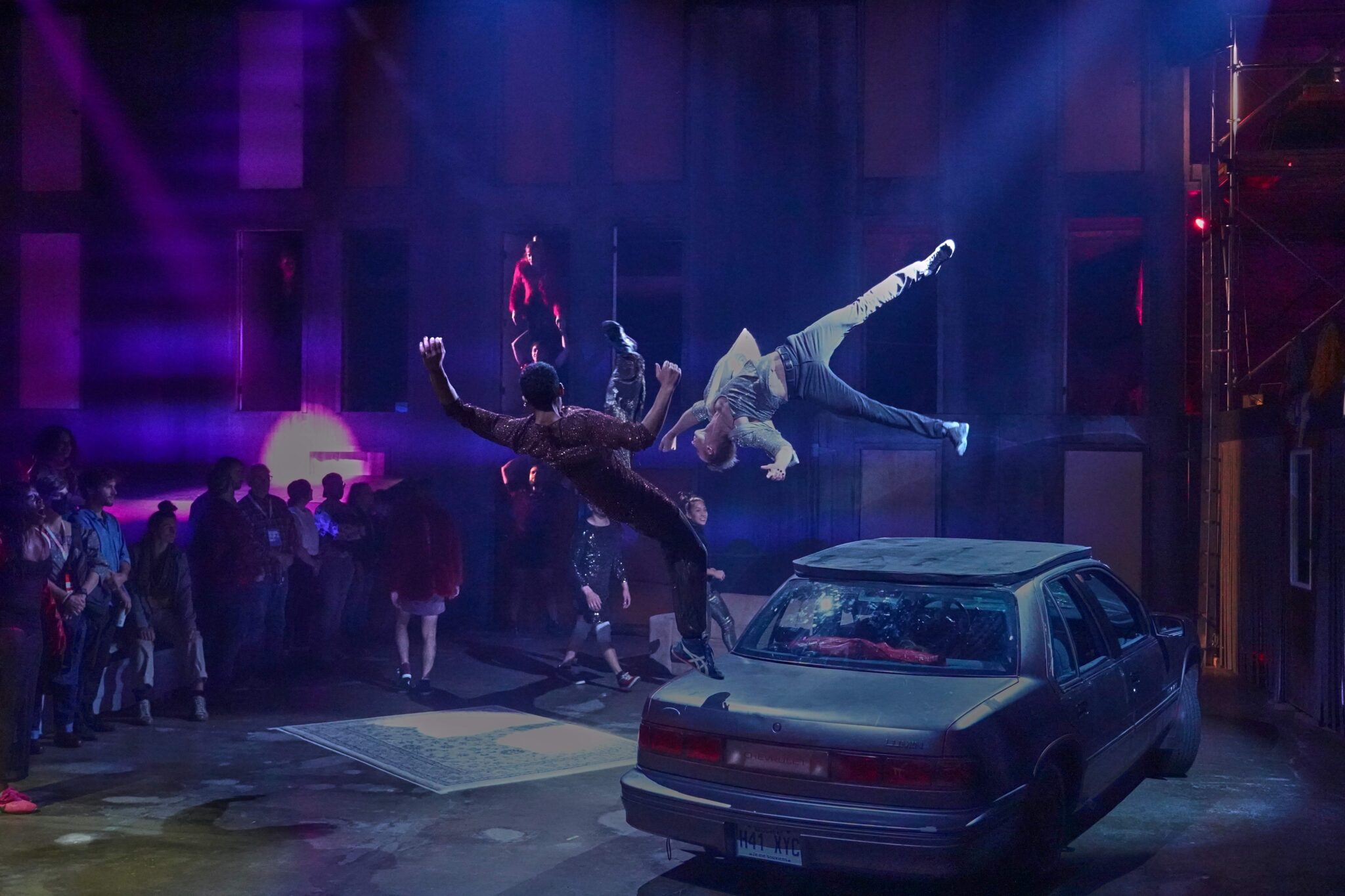 Deux danseurs en plein saut acrobatique au-dessus d'une voiture, sous des lumières violettes et devant un public