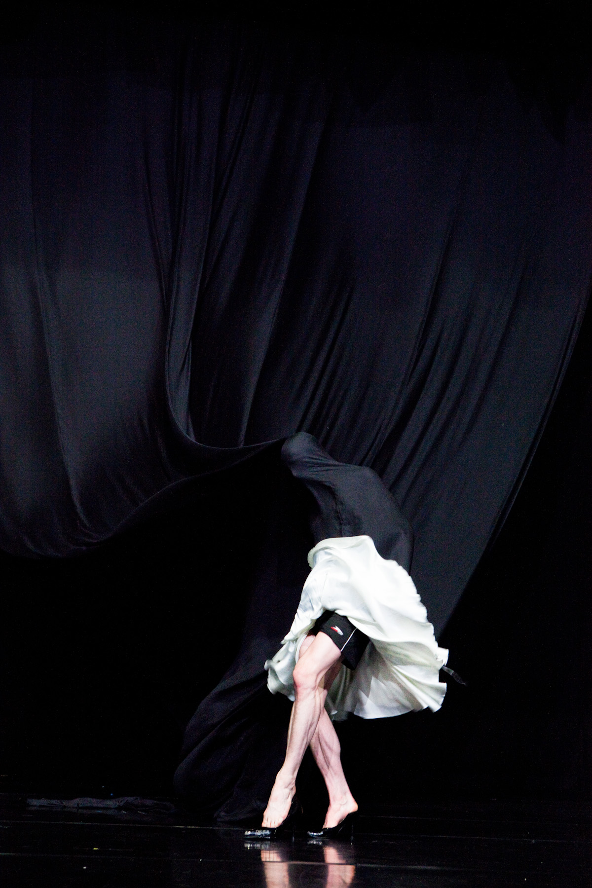 Un performeur sur scène jouant avec un tissu noir comme un rideau qui tombe du plafond, et dont la tête est cachée par ce même rideau, au cœur d’un dispositif scénique minimaliste.