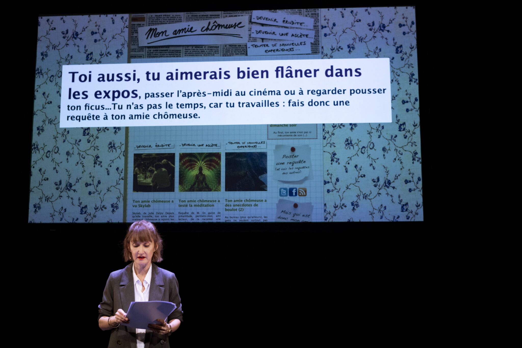 Sur scène, une femme lit un texte à haute voix, debout devant une projection humoristique titrée « Mon amie chômeuse » sur fond de motif fleuri.