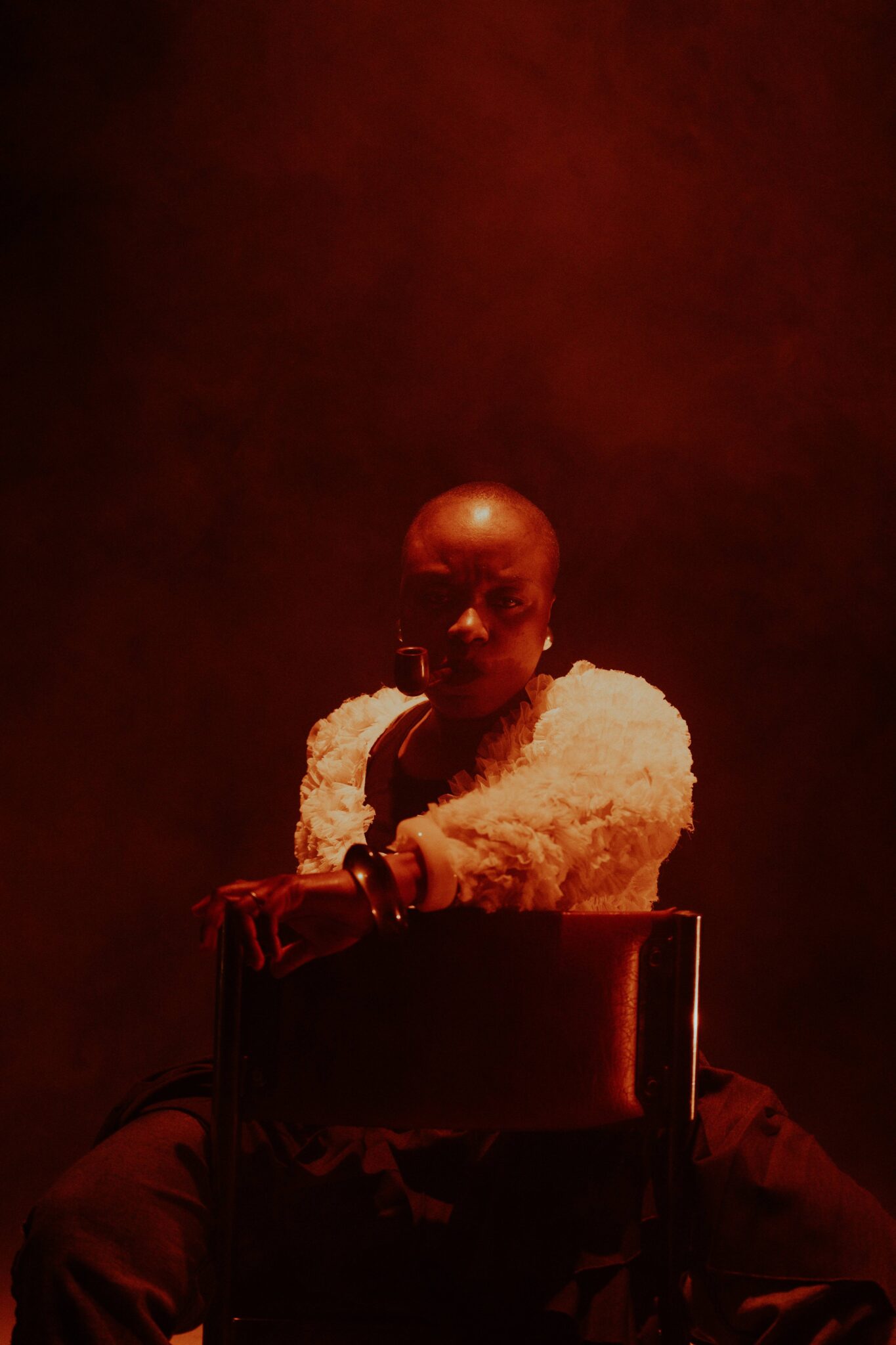 L’Effet mère, pièce de Carmel Loanga : une interprète assise de face, éclairée par une lumière rouge sombre, tenant une pipe et regardant le public.