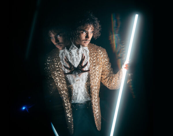 Un homme en veste dorée scintillante tient un tube lumineux blanc, son reflet multiplié de manière onirique dans une pénombre dramatique.