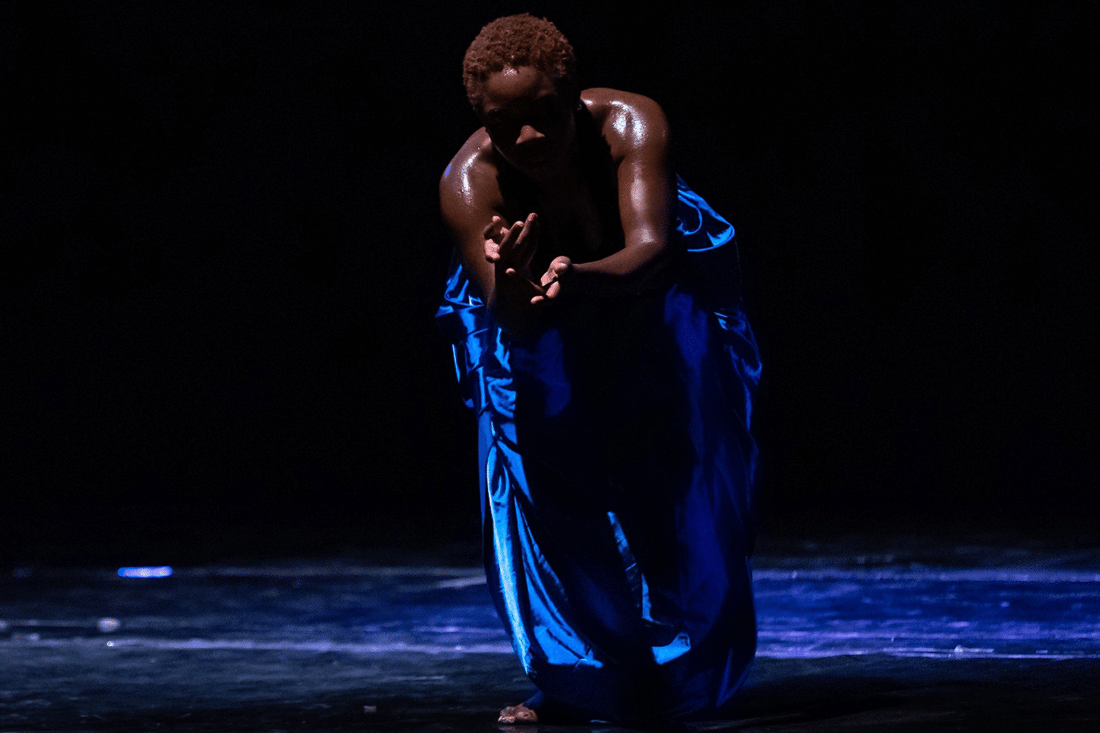 Nadia « Nadéeya » Gabrieli Kalati, chorégraphe et interprète du spectacle Sawata sur scène, en mouvement, éclairée par une lumière dramatique, vêtue d’un pantalon bleu et d’un haut noir.