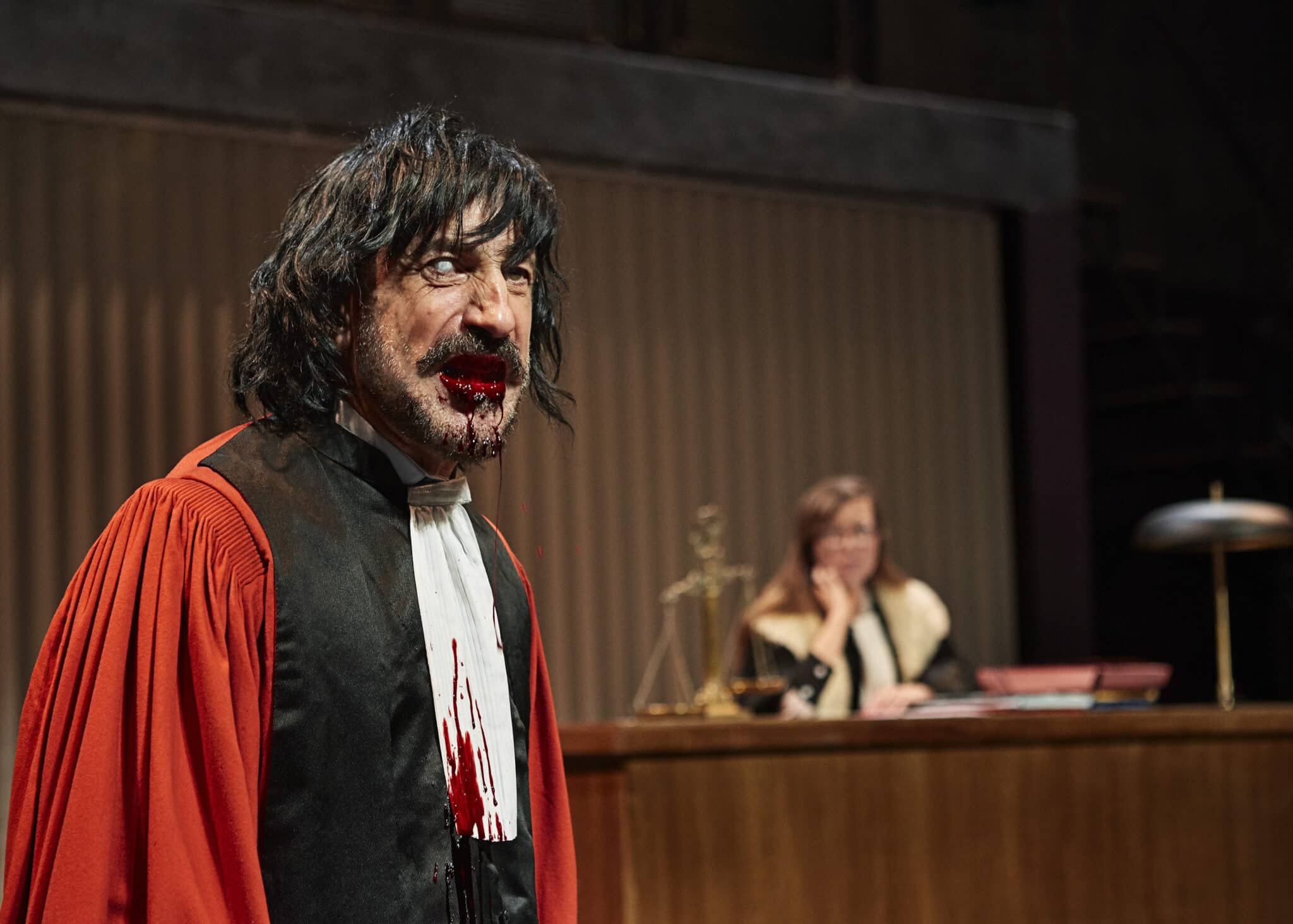 Un comédien des Chiens de Navarre, en costume sombre, s’exprime sur scène devant un décor évoquant un tribunal, dans le spectacle I will survive.