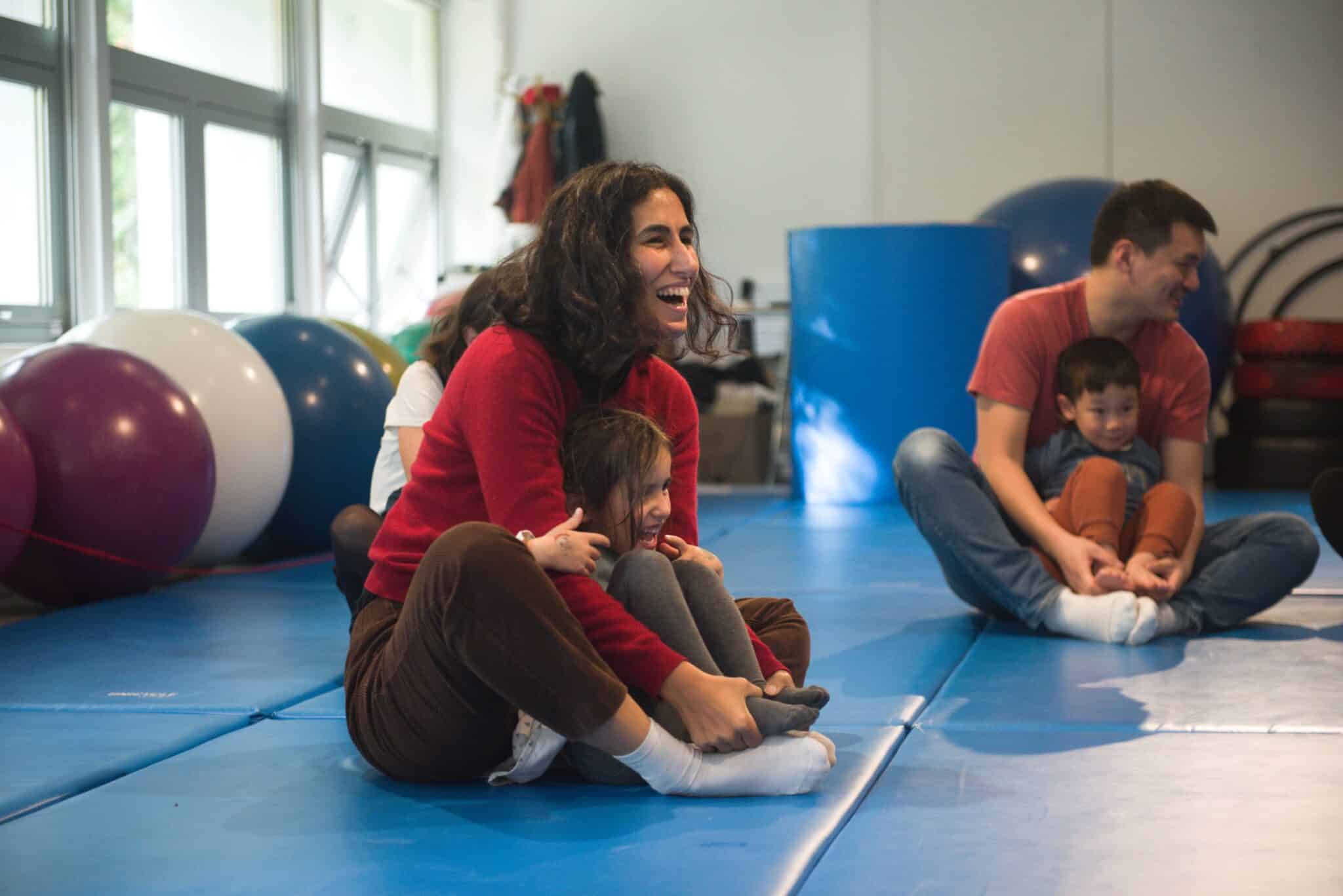 Adultes et enfants assis sur des tapis bleus, participant à un atelier Little Cirque dans une ambiance joyeuse.