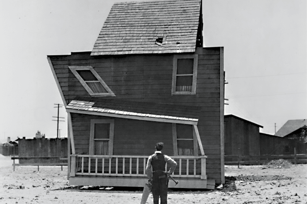 Homme en noir et blanc regardant une petite maison penchée et bancale.