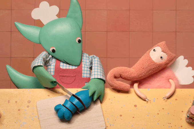 Personnage vert en tablier découpant un aliment bleu à côté d’une créature rose ailée.