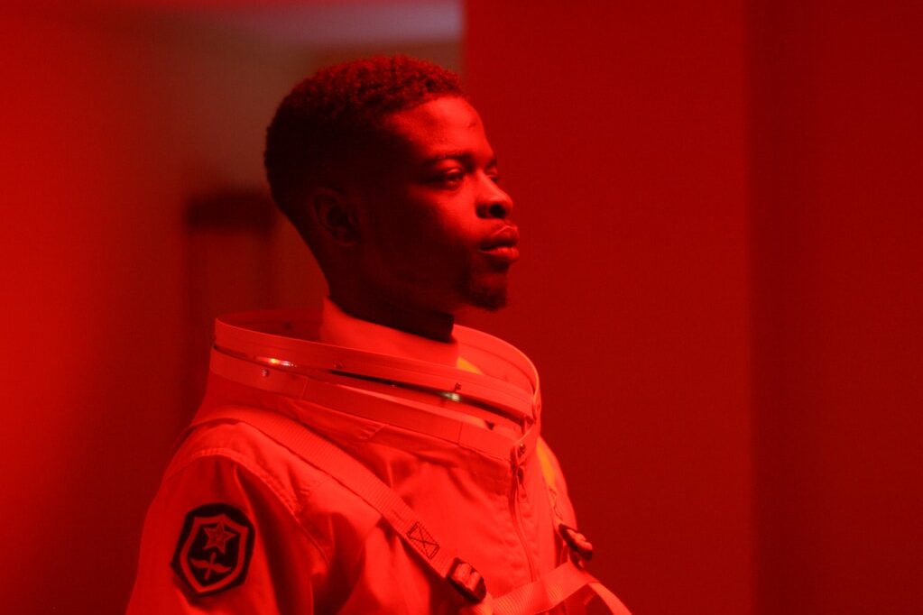 Jeune homme en tenue de cosmonaute dans une ambiance rouge. Son air est sérieux et déterminé.