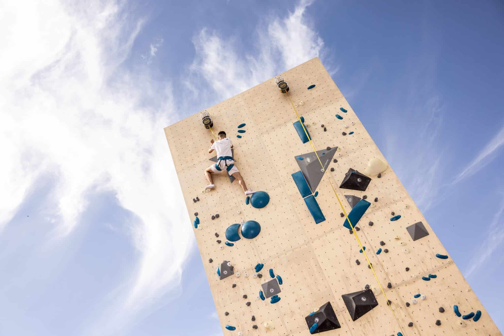 Personne pratiquant l’escalade sur un mur extérieur au Parc de la Villette sous un ciel bleu lors du projet Archi-Folies 2024, pendant les Jeux Olympiques 2024