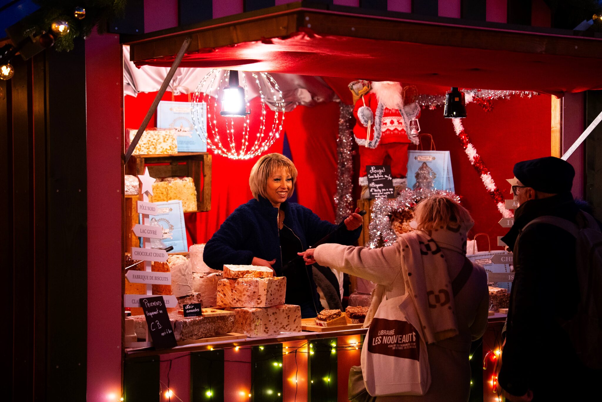 Stand de nougats décoré pour Noël, avec une vendeuse servant deux visiteurs.